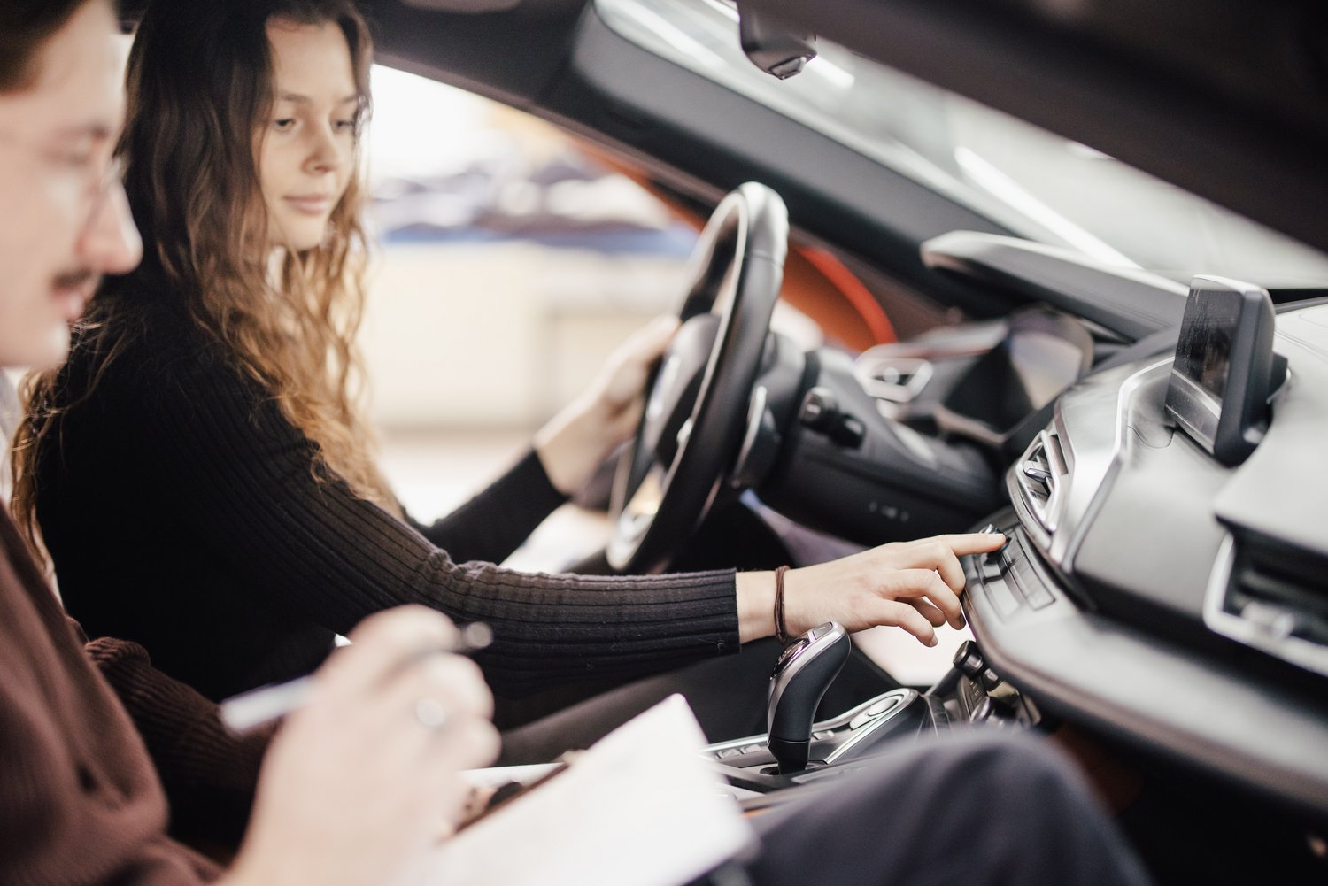 Eine Person sitzt am Steuer eines Autos und bedient das Armaturenbrett, während eine andere Person daneben Notizen macht. Beide konzentrieren sich auf die Bedienung und das Fahrzeuginnere.