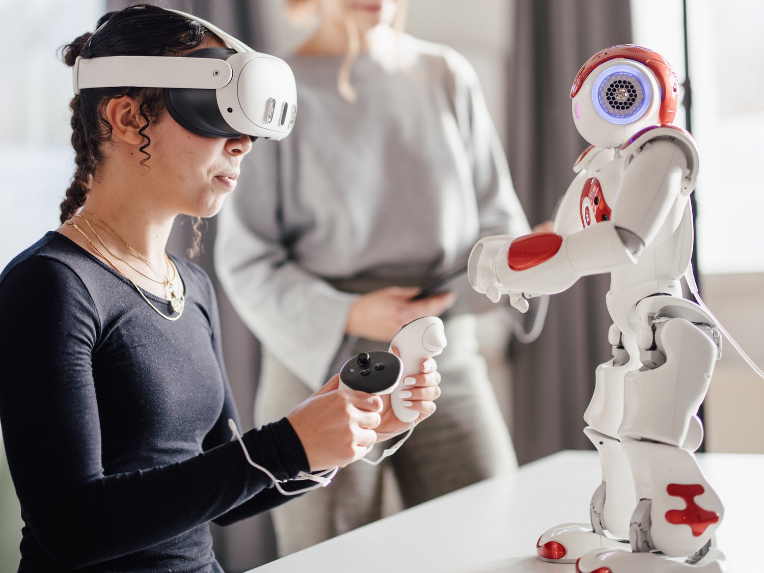 Eine Person mit VR-Brille sitzt an einem Tisch und hält einen Controller, während ein humanoider Roboter daneben steht. Eine weitere Person beobachtet die Interaktion im Hintergrund. Helle, moderne Umgebung.