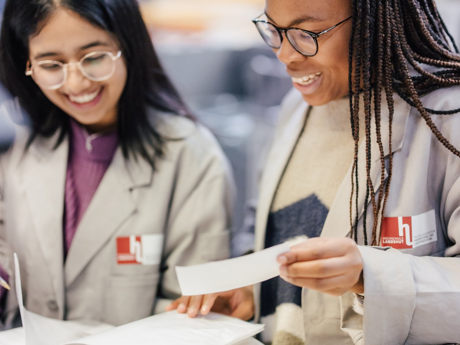 Zwei Personen in Laborkitteln stehen an einem Tisch und betrachten ein Buch. Sie lächeln und scheinen sich über den Inhalt auszutauschen, während im Hintergrund eine Werkstattumgebung zu sehen ist.