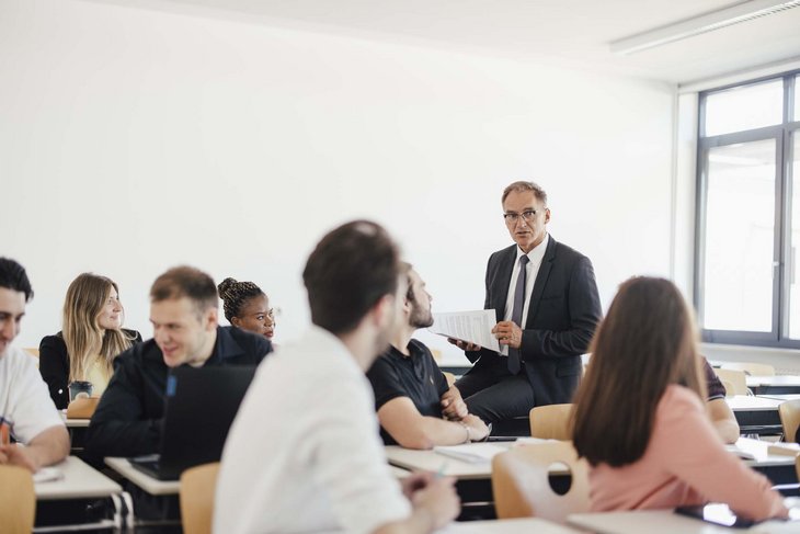 Mehrere Personen sitzen in einem Raum. Die meisten sitzen auf Stühlen nur einer sitzt auf einem Tisch und schaut die anderen an. Er hat Blätter in seiner Hand.