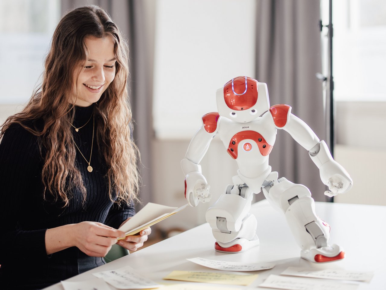 Eine Person sitzt an einem Tisch und betrachtet Karten, während ein humanoider Roboter daneben steht. Der Raum ist hell und modern, mit großen Fenstern und neutralen Farben.