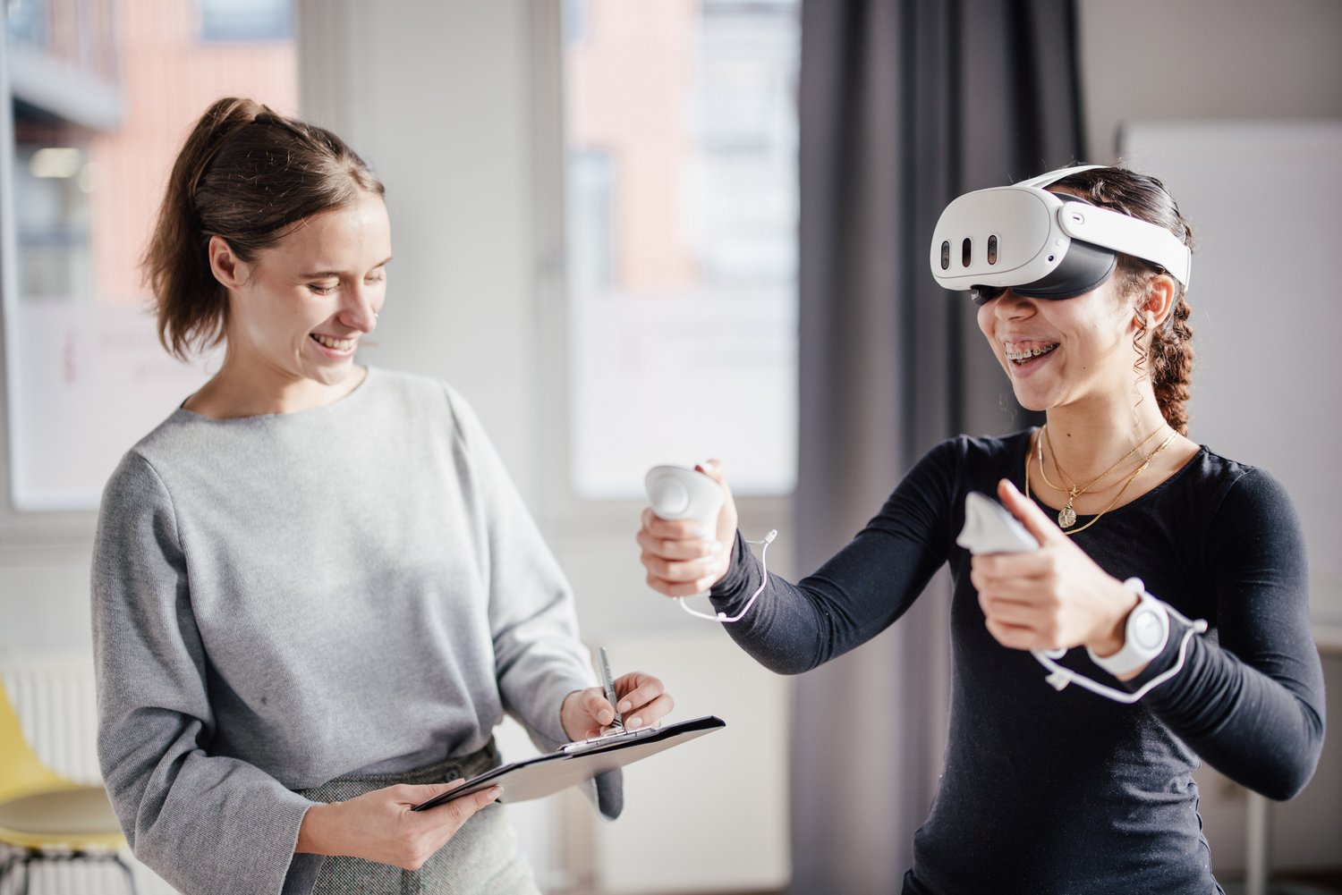 Eine Person trägt eine VR-Brille und hält Controller in den Händen, während sie lächelt. Eine andere Person steht daneben, notiert etwas auf einem Clipboard und beobachtet die Interaktion. Der Raum ist hell und modern eingerichtet.