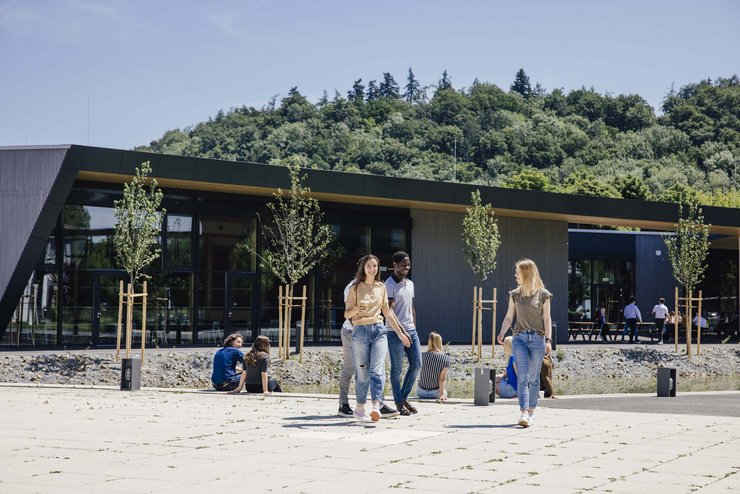 Man sieht Personen die draußen nebeneinander laufen, im Hintergrund ist das Gebäude der Mensa zu sehen.