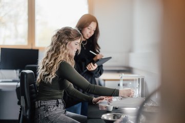 Zwei Personen arbeiten in einem Labor. Eine Person steht und hält ein Tablet, während die andere an einem Tisch mit einem Experiment beschäftigt ist. Helle, moderne Umgebung mit Fenstern im Hintergrund.