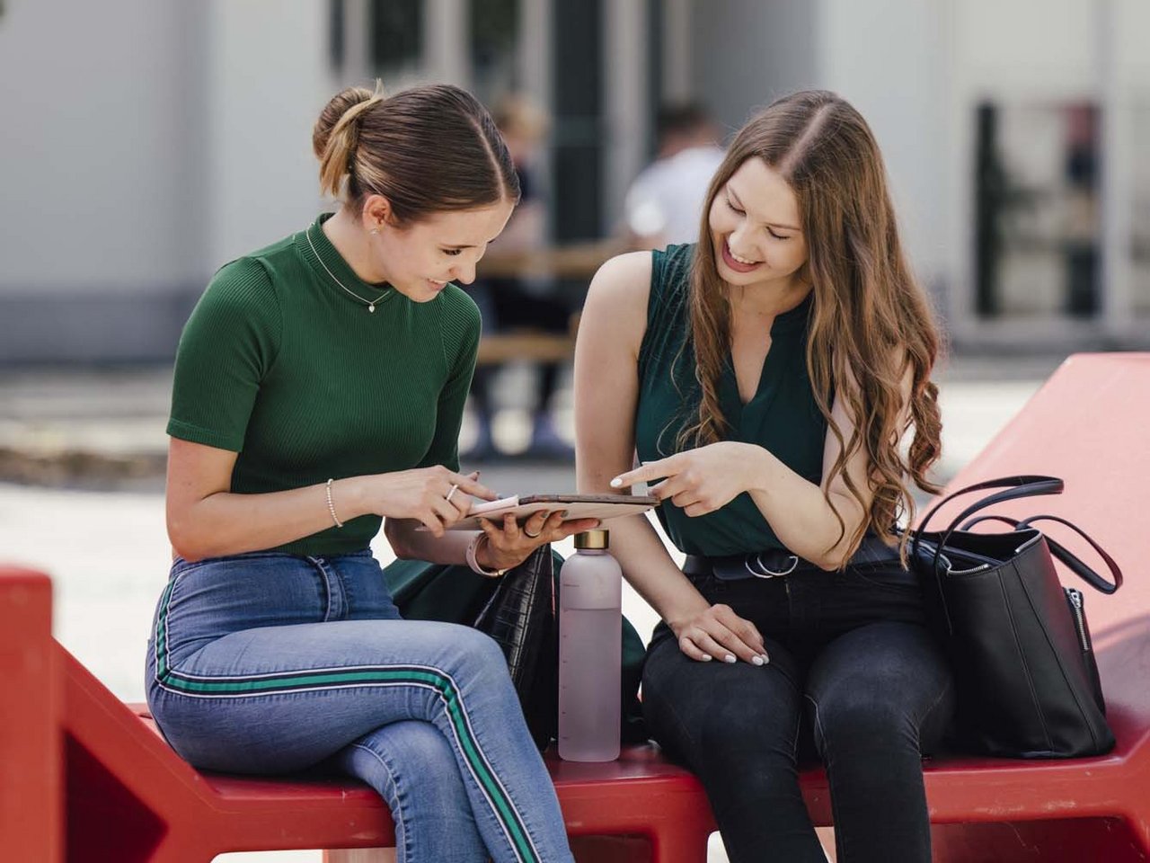 Zwei Personen sitzen auf einer Bank auf dem Campus. Eine der beiden hält ein Tablet. Beide schauen lächelnd in das Tablet rein.