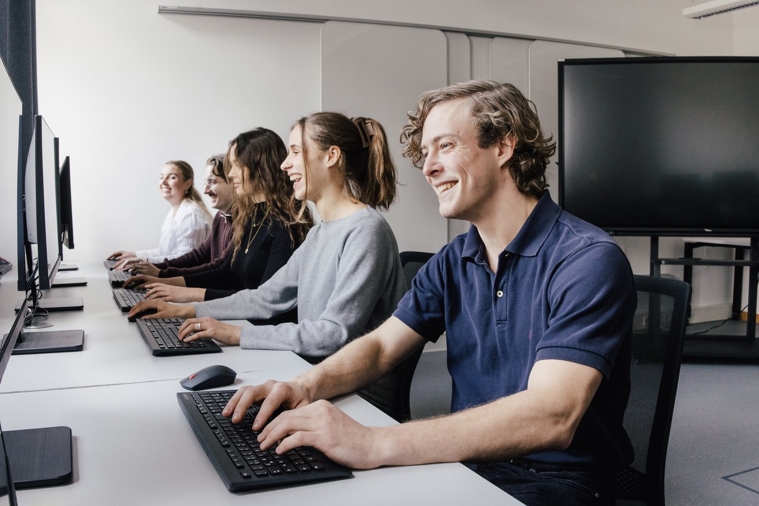 Sechs Personen sitzen an Computern in einem modernen Raum. Sie lächeln und arbeiten konzentriert an ihren Tastaturen. Der Raum ist hell und freundlich gestaltet, mit großen Bildschirmen im Hintergrund.