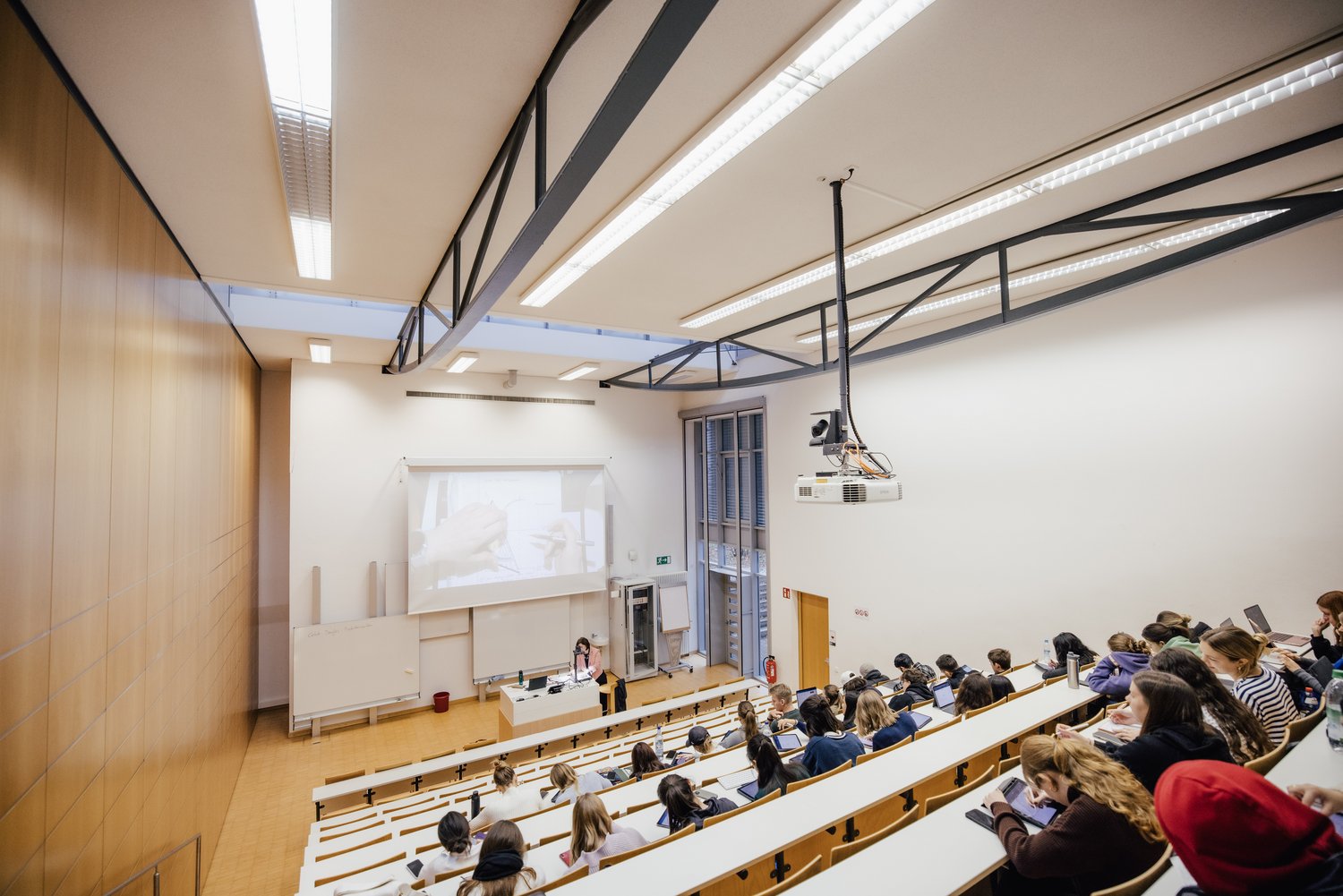 Ein großer Hörsaal mit mehreren Reihen von Sitzplätzen. Eine Person steht vorne und präsentiert an einer Leinwand, während die Zuhörenden aufmerksam auf ihre Geräte schauen. Helle, moderne Architektur mit viel Tageslicht.