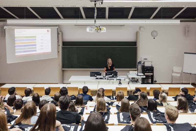 Man sieht mehrere sitzende Personen und eine Person die vorne im Raum an einer Tafel steht.