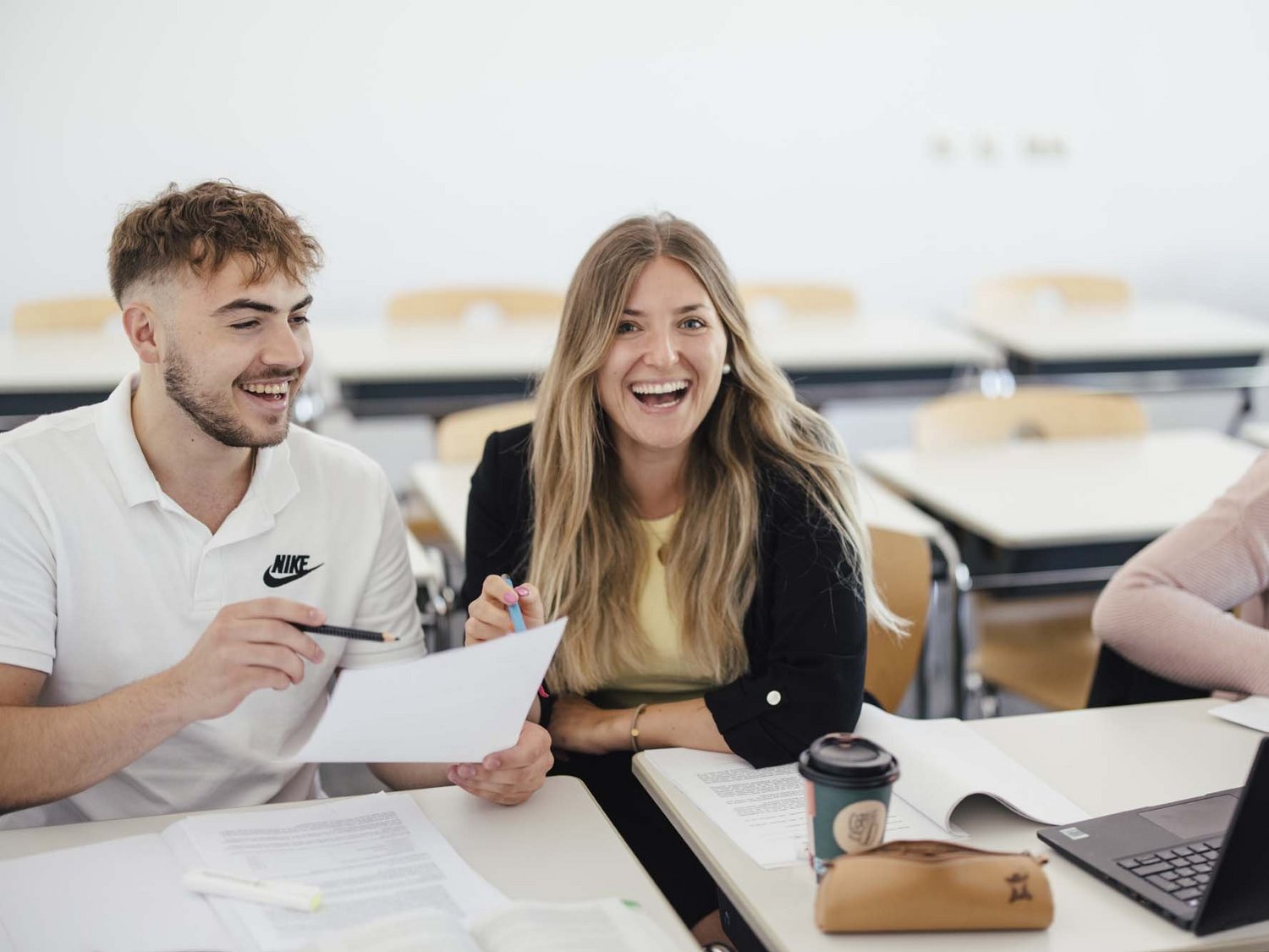 Zwei Personen sitzen in einem Raum beide freuen sich. Sie schauen zusammen in ein Blatt rein. Beide halten einen Stift in ihrer Hand