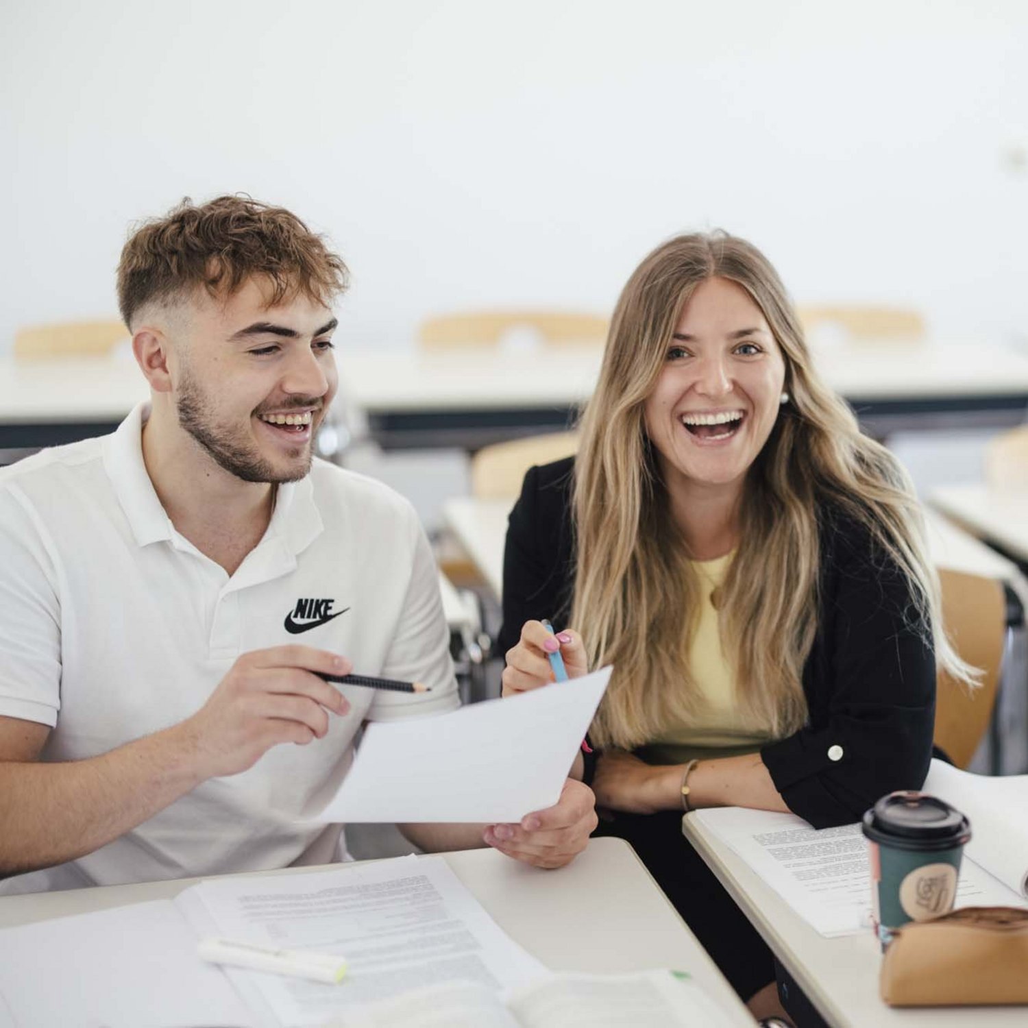 Zwei Personen sitzen in einem Raum beide freuen sich. Sie schauen zusammen in ein Blatt rein. Beide halten einen Stift in ihrer Hand