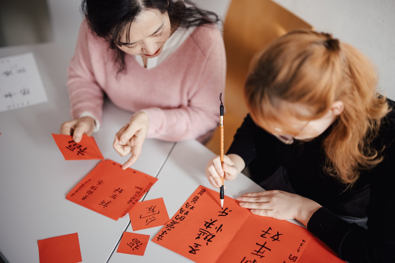 Zwei Personen sitzen an einem Tisch und schreiben mit einem Pinsel auf rote Papierstücke. Die Umgebung ist hell und freundlich, und die Schriftzeichen sind in traditionellem Stil gehalten.
