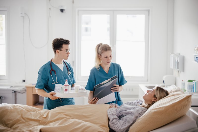 Ein medizinisches Team in blauen Scrubs steht neben einem Bett. Eine Person hält eine Tablett mit Medikamenten, während die andere eine Patientenakte zeigt. Eine Patientin liegt im Bett und hört aufmerksam zu.