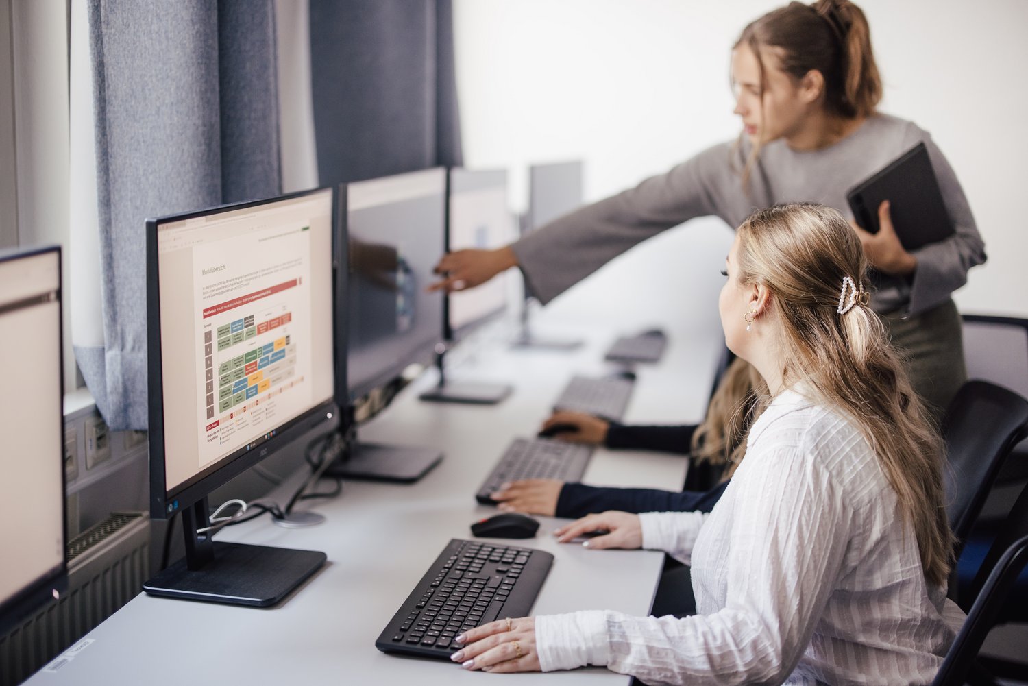 Zwei Personen arbeiten an Computern in einem modernen Raum. Eine Person zeigt auf den Bildschirm, während die andere konzentriert tippt. Der Raum ist hell und mit großen Fenstern ausgestattet.