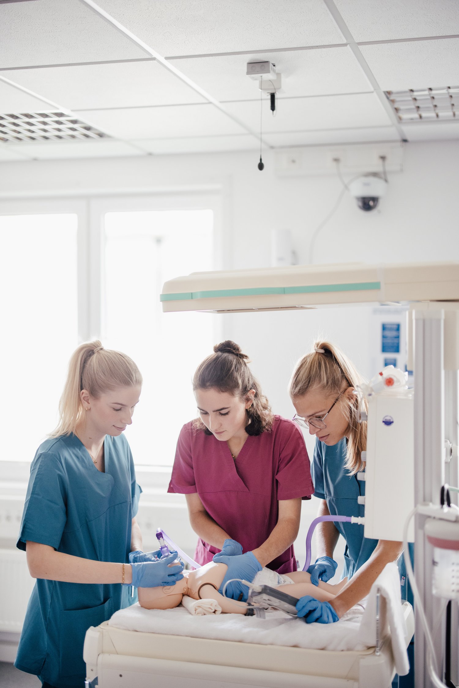 Drei Fachkräfte in medizinischer Kleidung arbeiten an einem Dummy in einem Krankenhaus. Eine Person führt eine Injektion durch, während die anderen assistieren. Der Raum ist hell und modern ausgestattet.