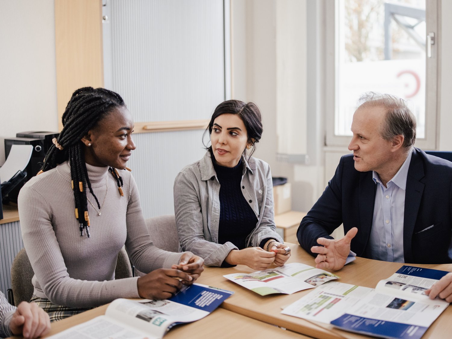Drei Personen sitzen an einem Tisch in einem Büro und diskutieren. Auf dem Tisch liegen Broschüren. Eine Person hat geflochtene Haare, die anderen haben unterschiedliche Frisuren. Alle wirken engagiert und interessiert.